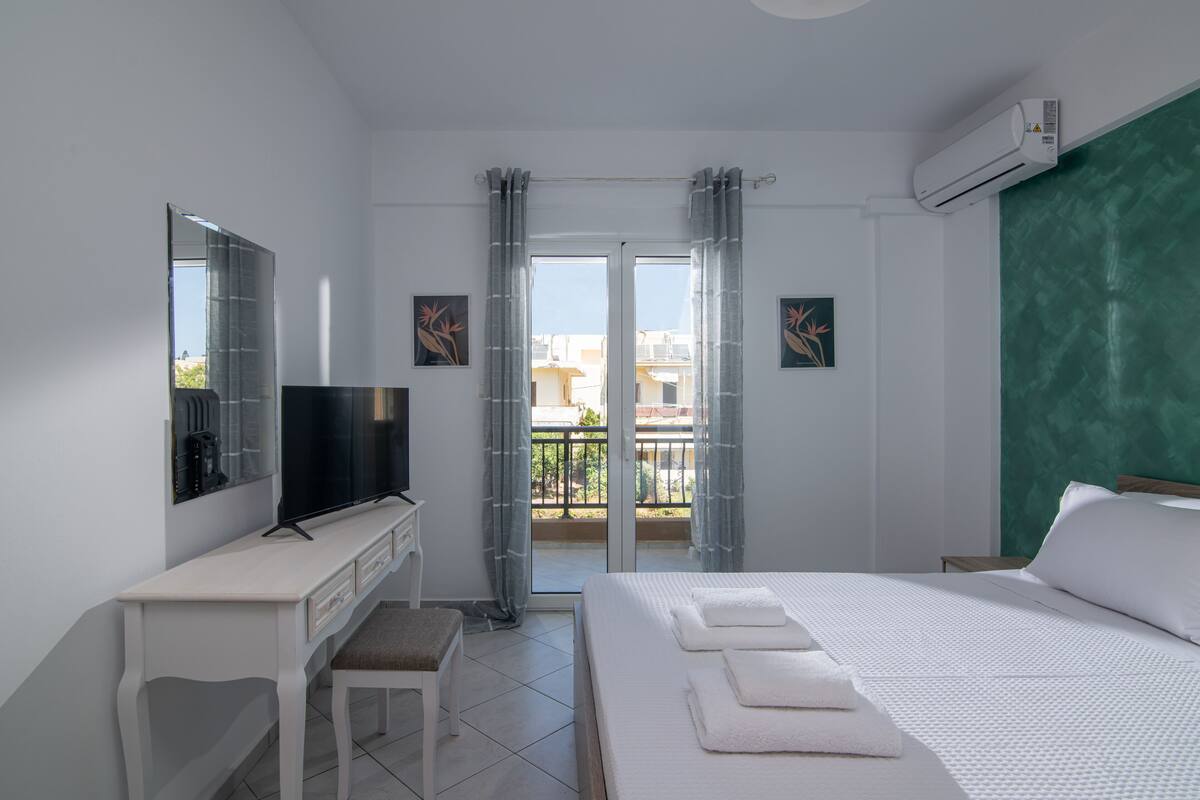 A hotel room with a bed, a desk with a TV, a balcony with curtains, and a green accent wall.