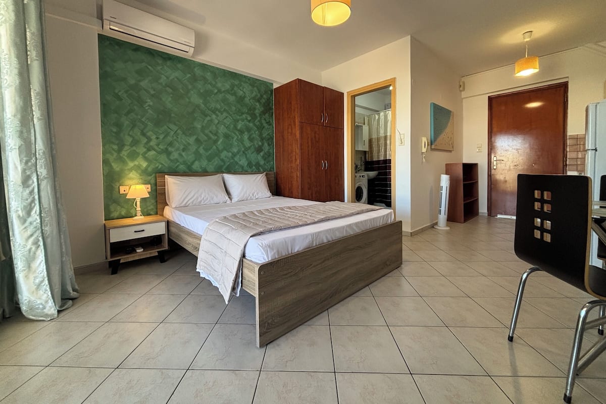 A bedroom with a bed, a nightstand, an air conditioner, a wardrobe, a desk with a chair, and a bathroom visible through an open door.