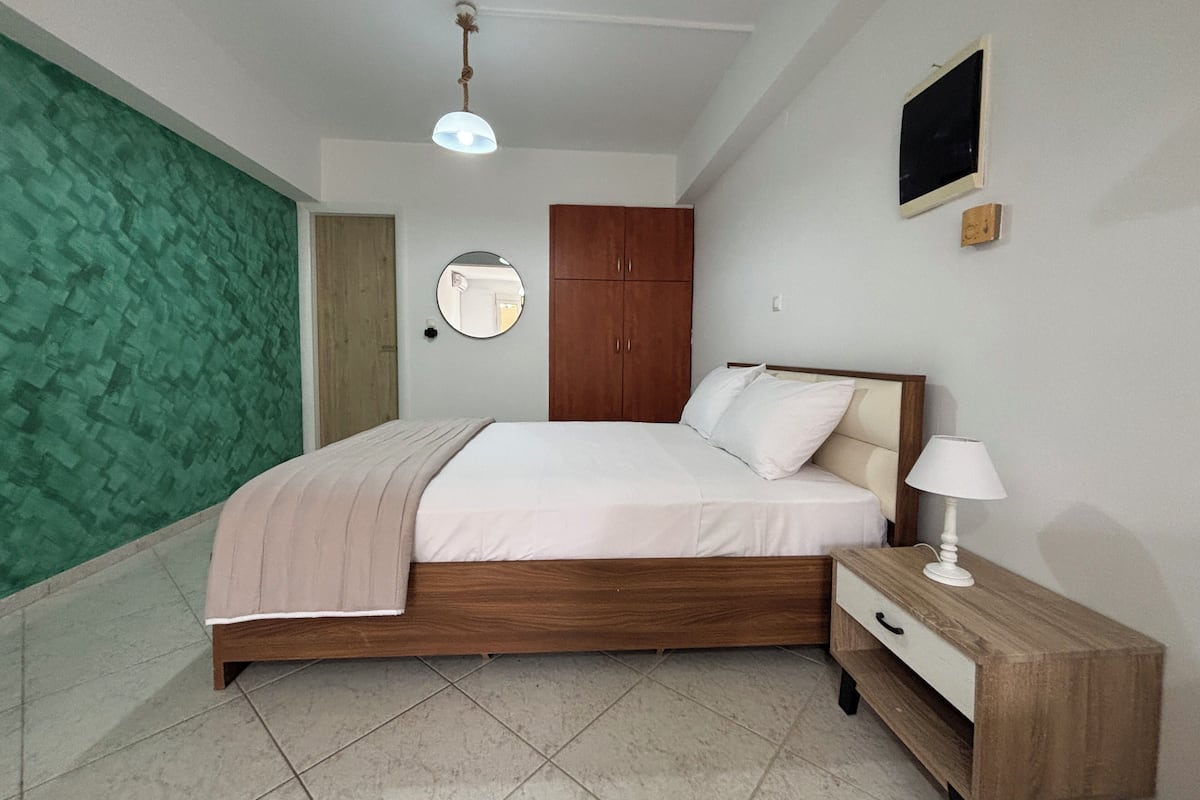 A bedroom with a bed, bedside table, lamp, wardrobe, mirror, and a green textured wall.