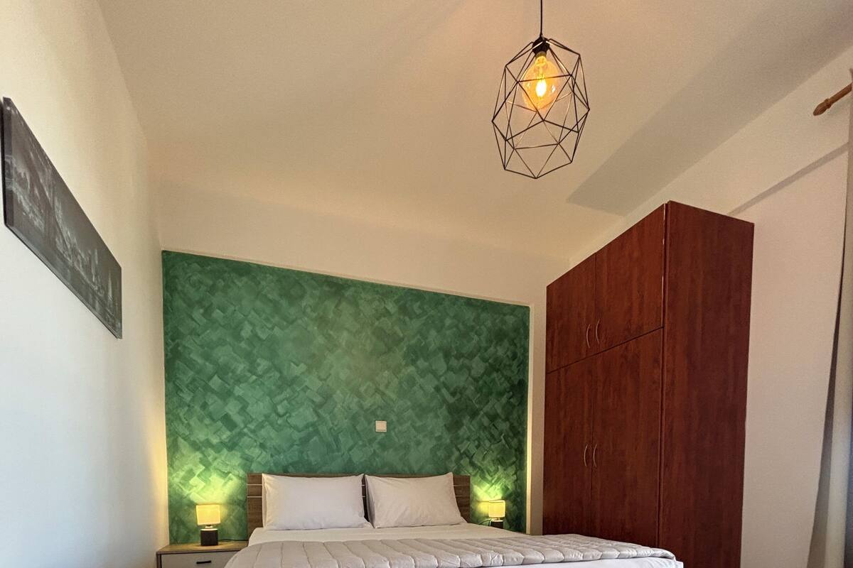 A bedroom with a bed, a green textured wall, a wooden wardrobe, and a hanging light fixture.