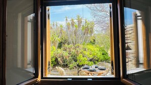 Une vue depuis une fenêtre à cadre en bois, donnant sur un jardin où l’on peut voir différentes plantes et une table mise pour le repas.