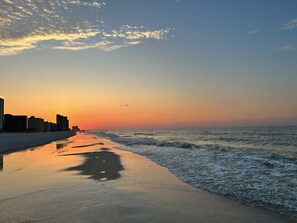 Uma praia com um pôr do sol, ondas e edifícios ao fundo.