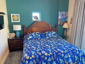 A bedroom with a wooden headboard, a bed with a blue floral bedspread, a nightstand with a lamp, and a painting on the wall.