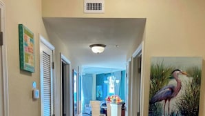 A hallway with beige walls, a tiled floor, and a painting of a bird. There are doors on the left, a window with curtains, and a dining area in the background. - Crystal Towers 609, Corner Unit
Your Perfect Beach Getaway Awaits You! (Gulf Shores)