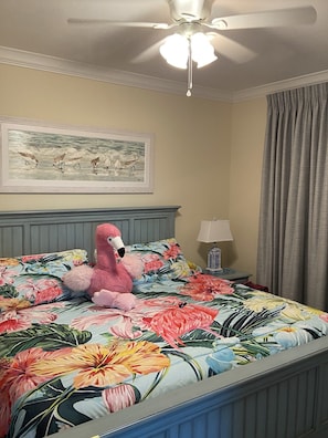 Quarto com colcha floral, ventilador de teto, pintura na parede e abajur em uma mesinha de cabeceira.
