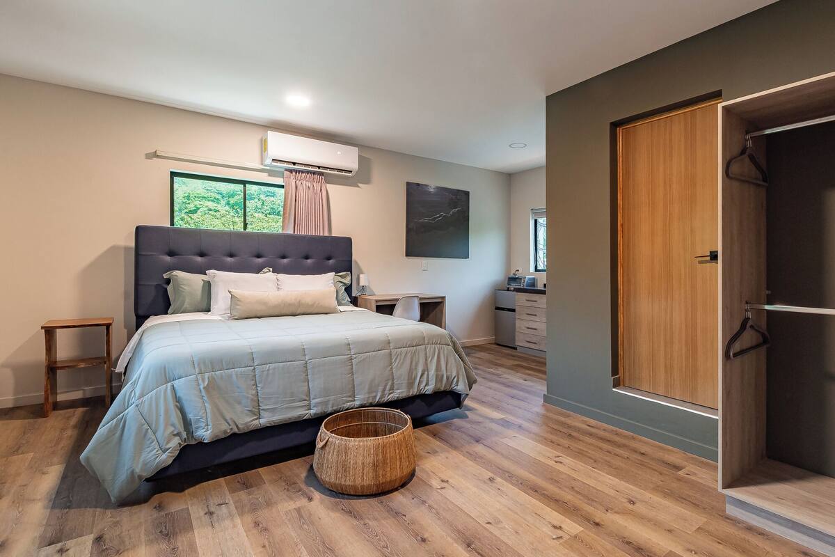 Quarto com cama, mesas de cabeceira, janela com cortinas, ar condicionado, porta de madeira e uma pequena cesta no chão.