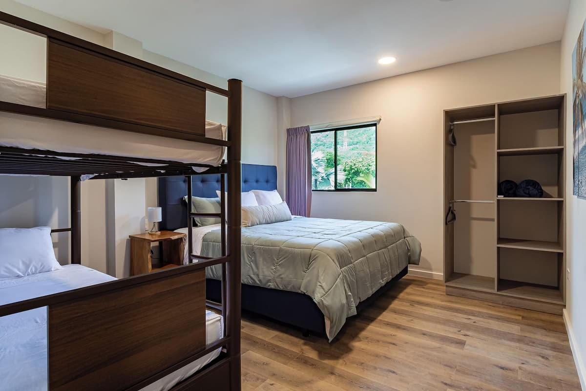 Quarto com uma cama beliche, uma cama de solteiro, mesa de cabeceira com abajur, janela com cortinas e um closet espaçoso.
