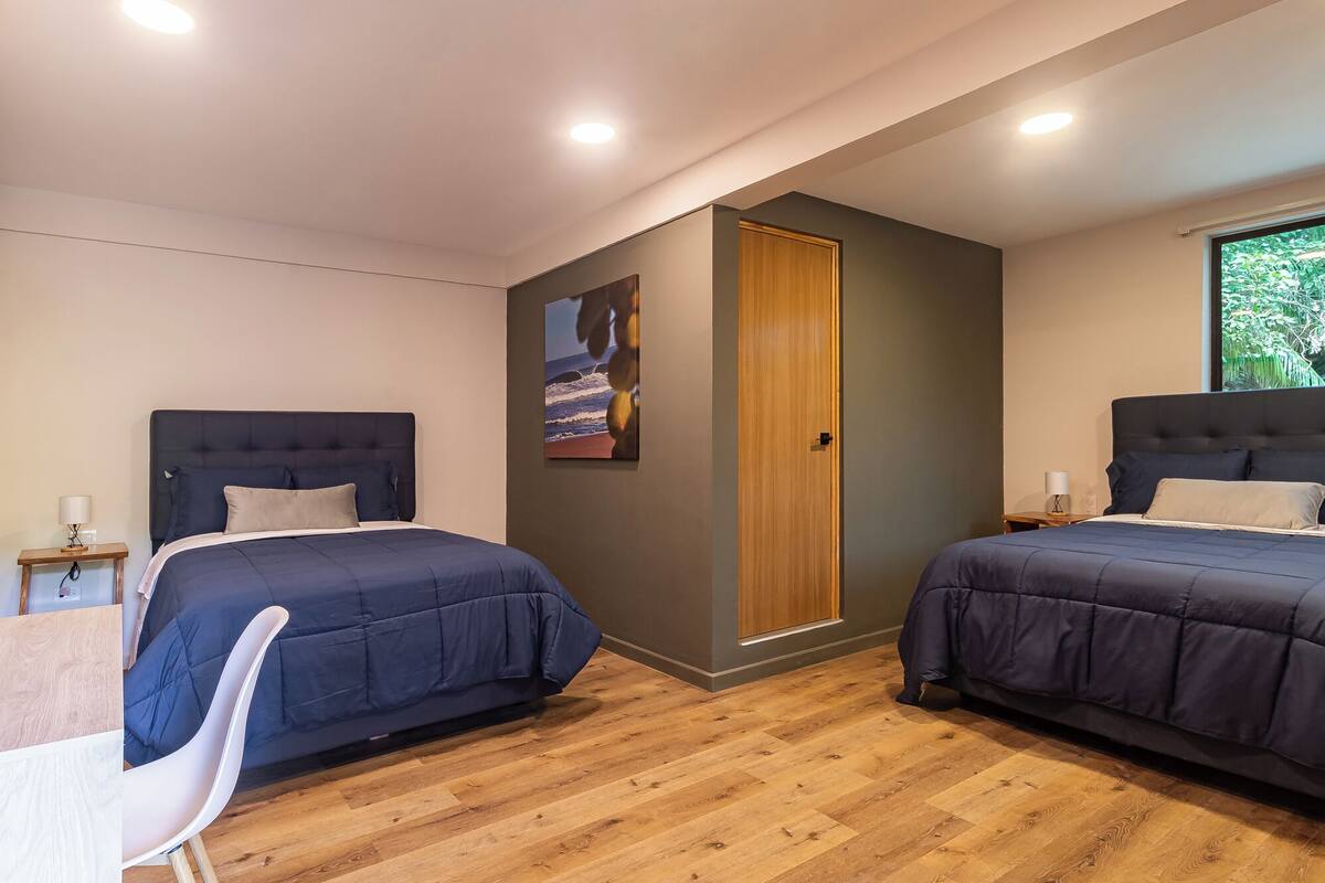 Quarto moderno com duas camas, piso de madeira, porta de madeira e janela com vista.