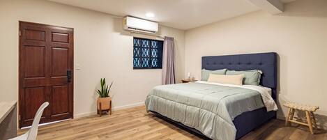 A bedroom with a wooden floor, a bed with a blue headboard, a potted plant, a desk, and a door.
