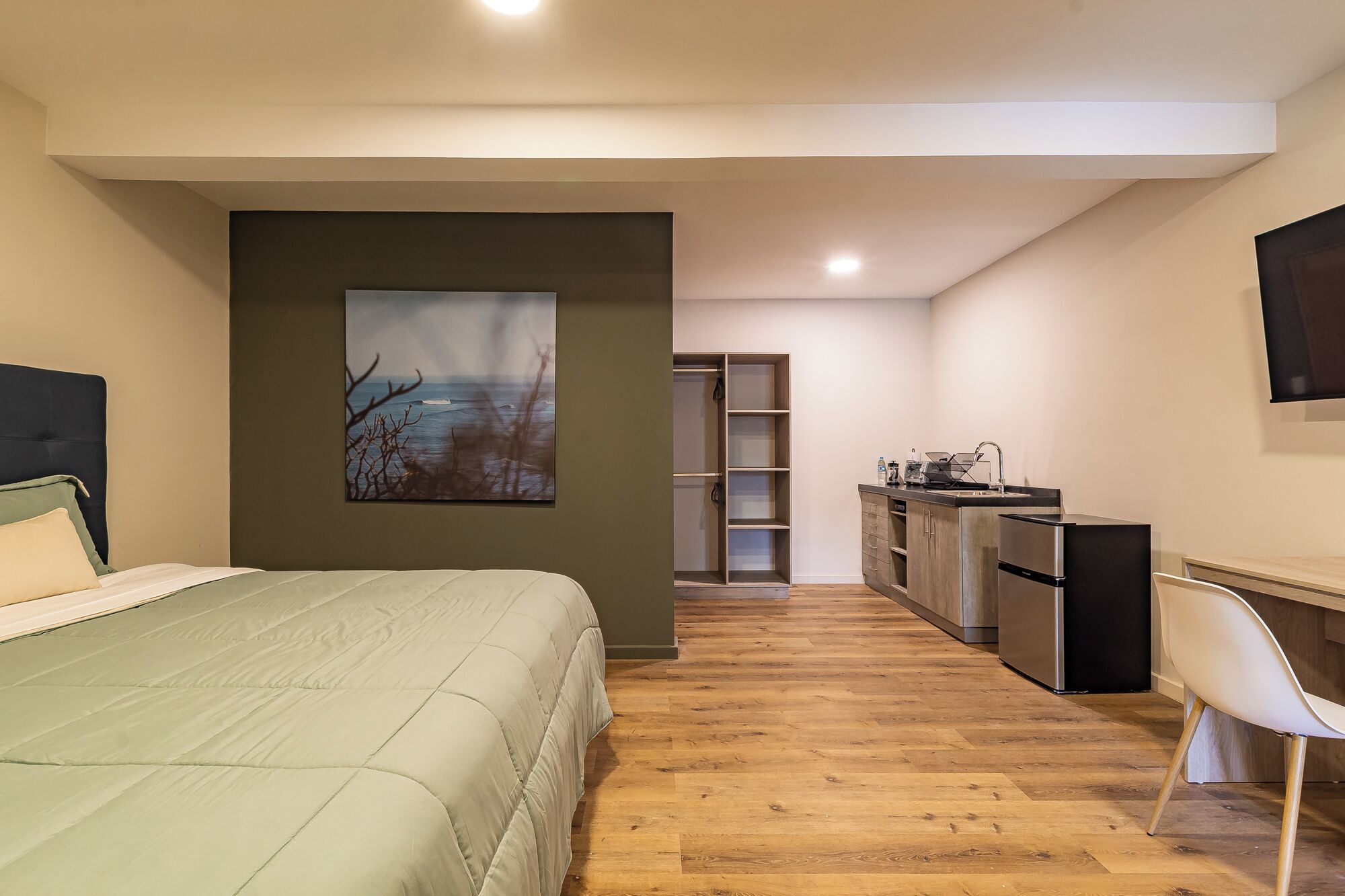 A bedroom with a bed, a painting, a kitchenette, and a chair.