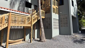 A modern building with a wooden staircase leading to a balcony, surrounded by palm trees and a gravel ground. - Lora Loca Inn (El Zonte)