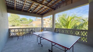 A covered outdoor area with a ping pong table, chairs, and a view of palm trees and buildings. - Lora Loca Inn (El Zonte)