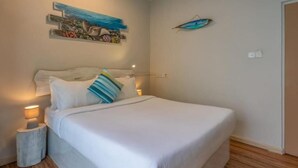 Un lit bien fait, avec une literie blanche et un coussin à rayures, une petite table de chevet avec une lampe, et une décoration murale représentant un poisson et des fleurs.
