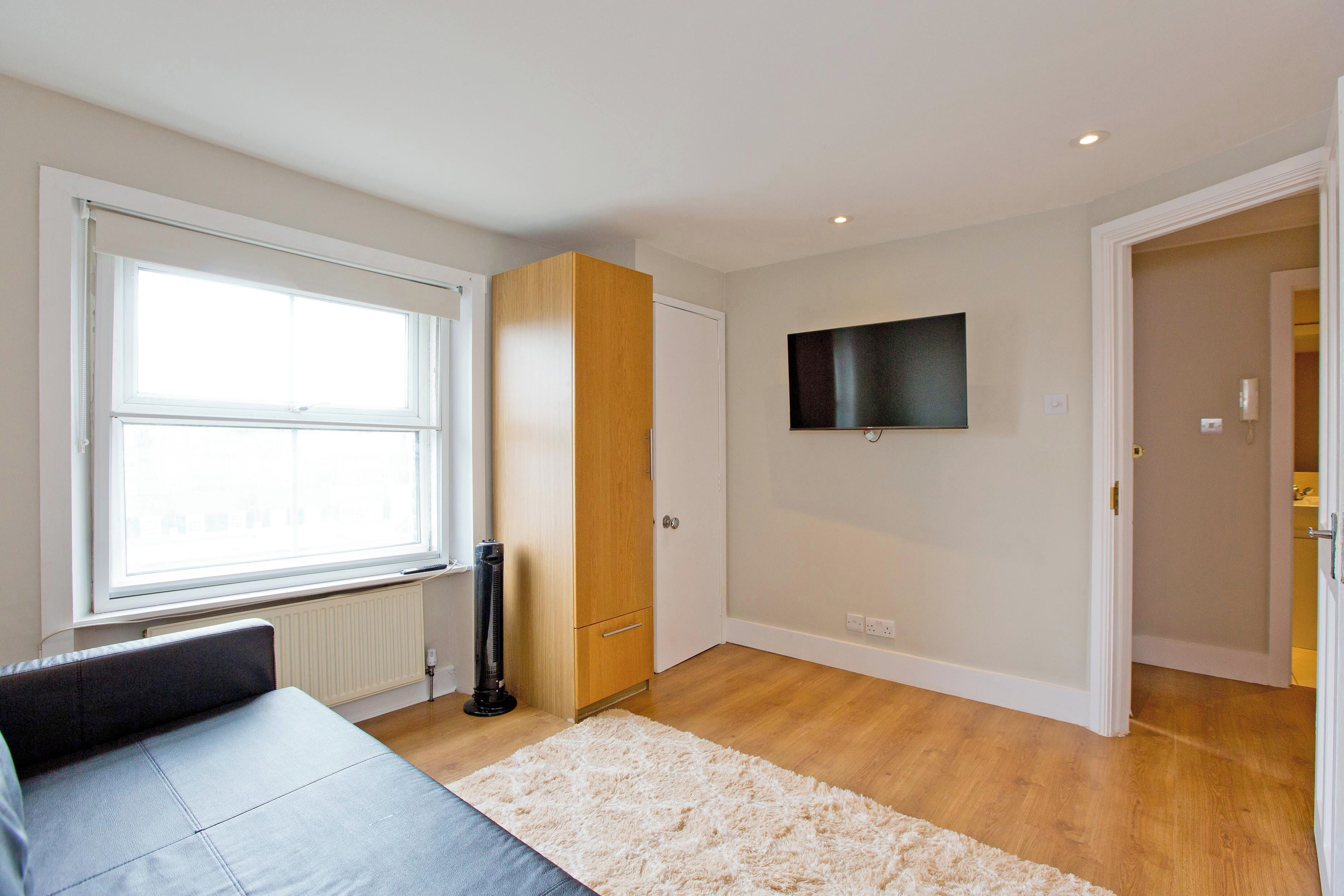 A modern room with a large window, a flat-screen TV mounted on the wall, a wooden wardrobe, and a white carpet.