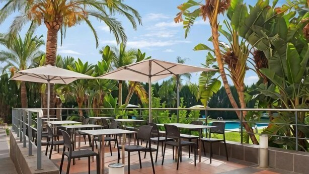 Um pátio externo com mesas, cadeiras, guarda-sóis e uma piscina ao fundo.