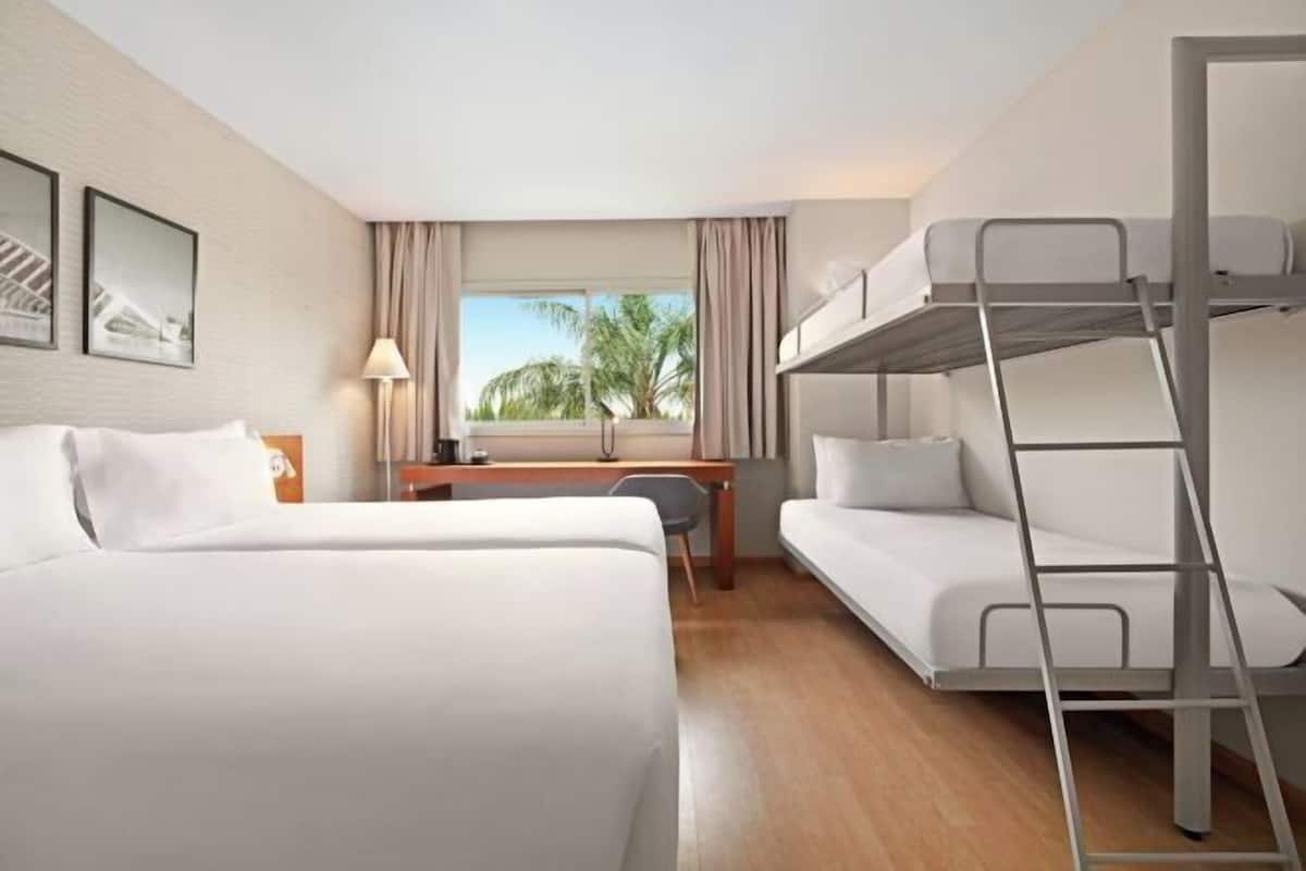 A hotel room with two beds, a desk, a chair, a bunk bed, and a window with a view of palm trees.