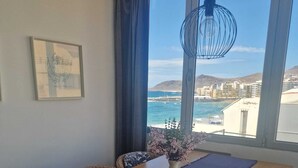 A dining area with a long table, wicker chairs, a vase with flowers, and a view of the sea and buildings. - Ocean Terrace Quarries. (Las Palmas de Gran Canaria)