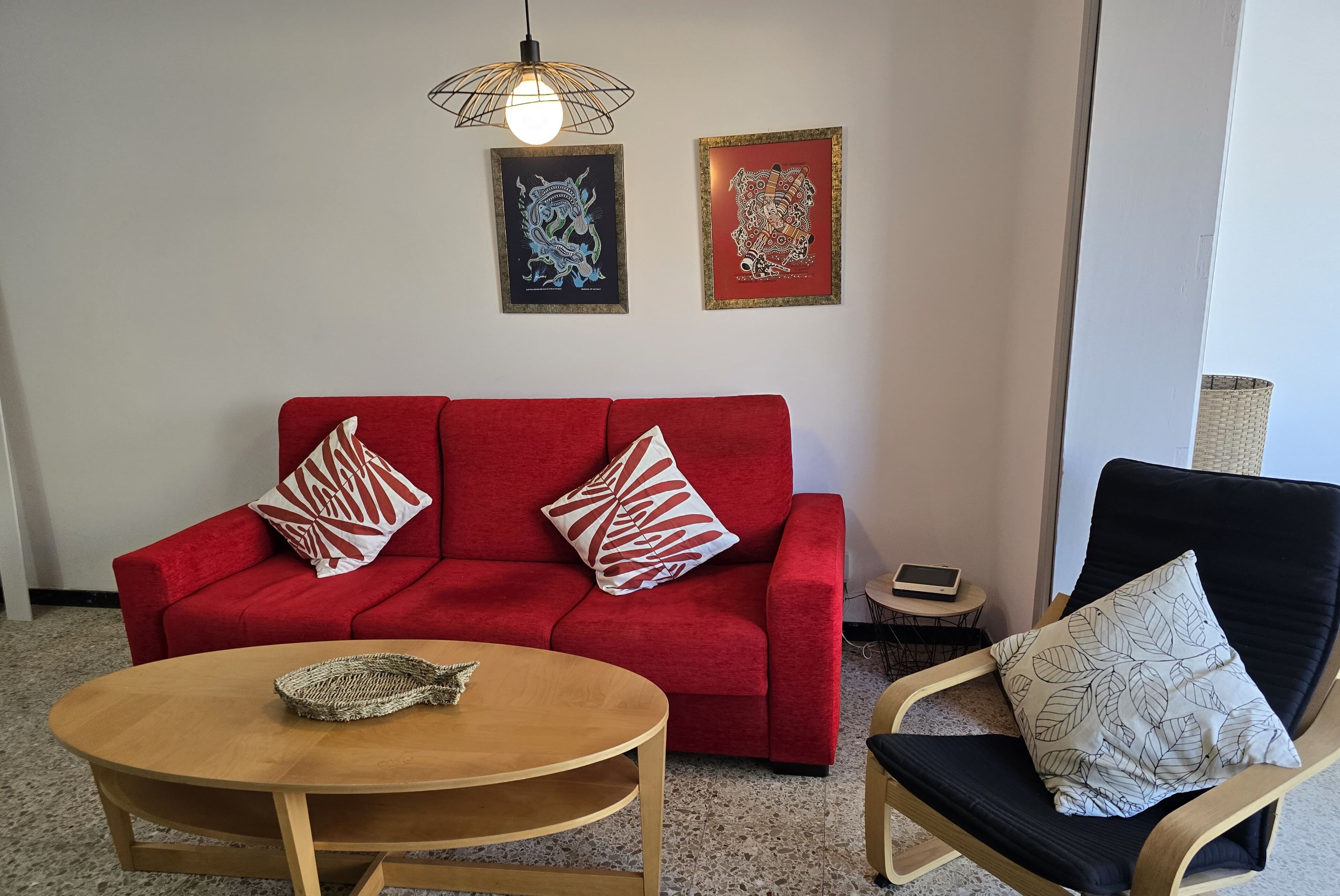 En rød sofa med mønstrede puder, et træbord og en sort lænestol med pude i bladmotiv. To billeder hænger i væggen over.