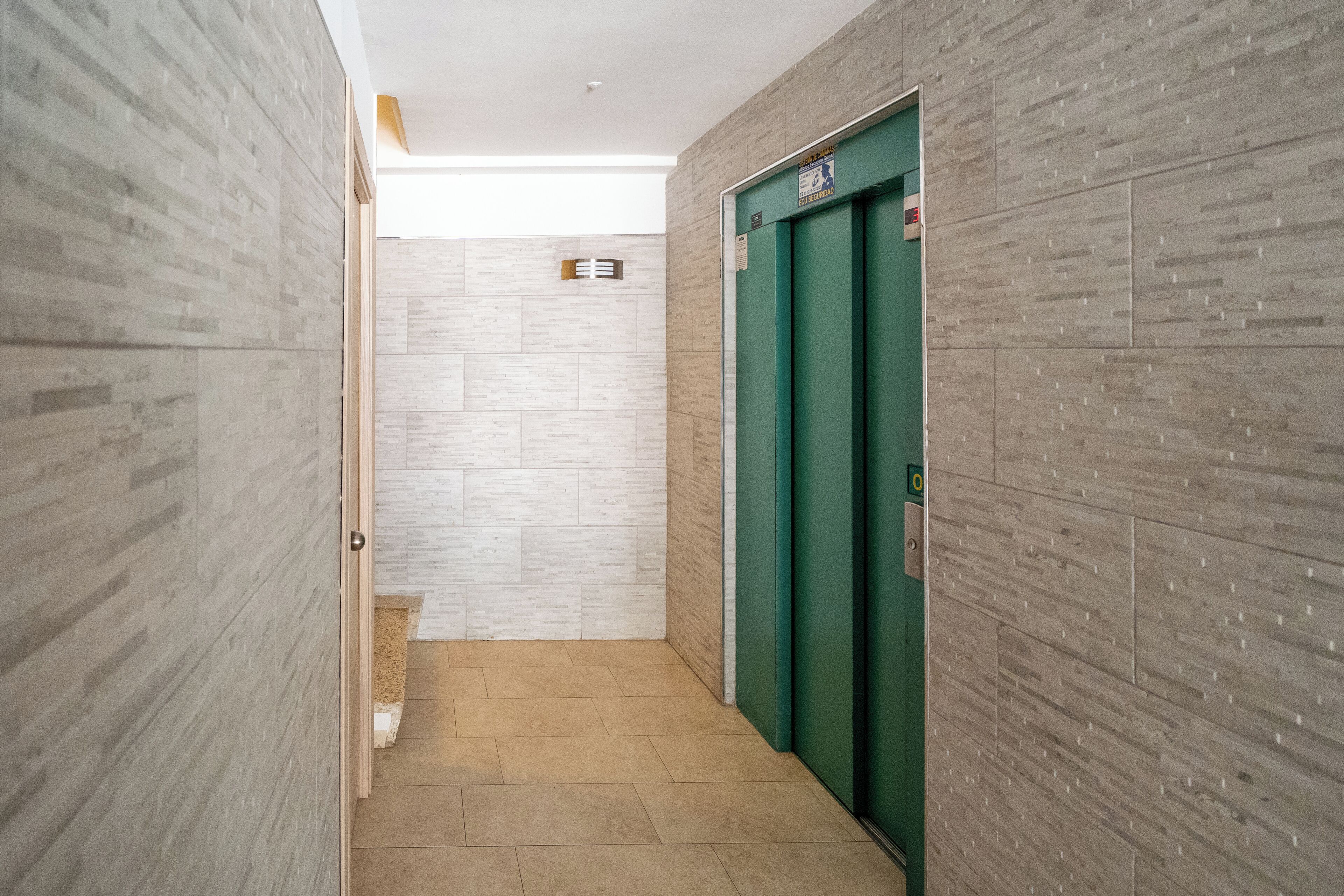 A narrow hallway with a tiled floor and walls, leading to a green elevator.