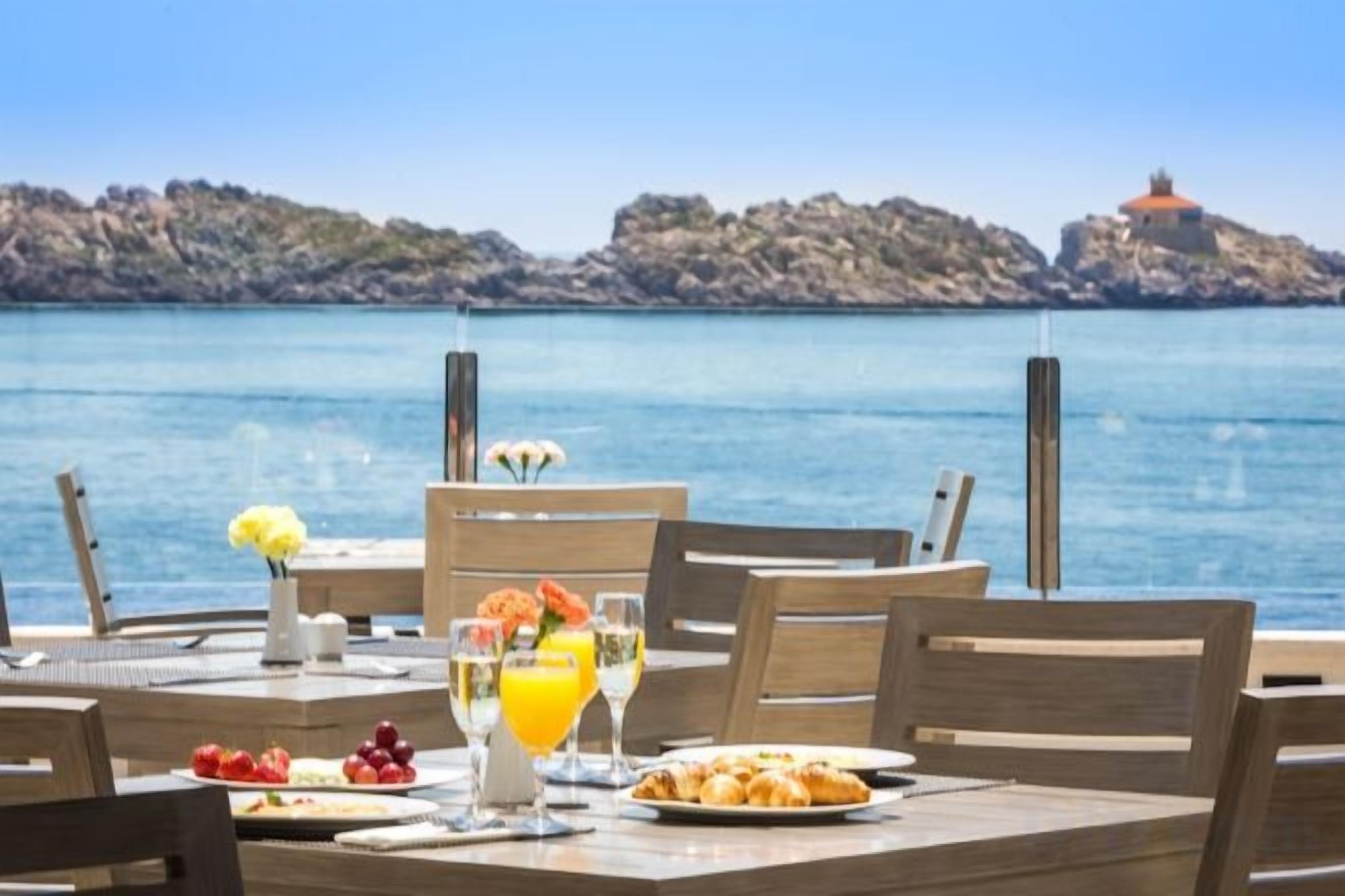 A dining table set with breakfast items and a glass of orange juice, overlooking a body of water with a rocky shoreline.