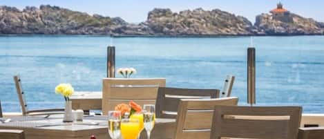 Una mesa de comedor dispuesta con alimentos para el desayuno y un vaso de jugo de naranja, con vista a un cuerpo de agua y una costa rocosa.
