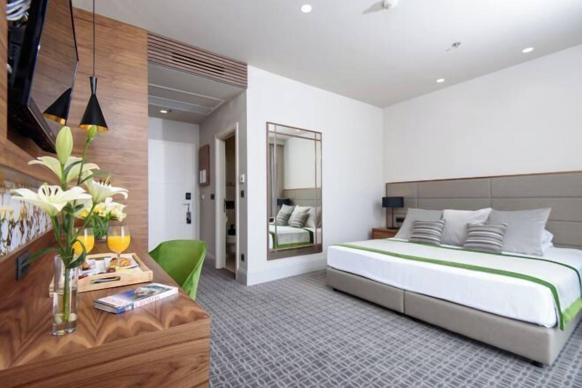 Una habitación de hotel moderna con una cama grande, un escritorio de madera con un florero y un espejo, y una silla verde.