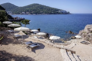 A beach with sun loungers and umbrellas, rocky shoreline, and a view of a distant hill. - Royal Neptun Hotel (Dubrovnik)