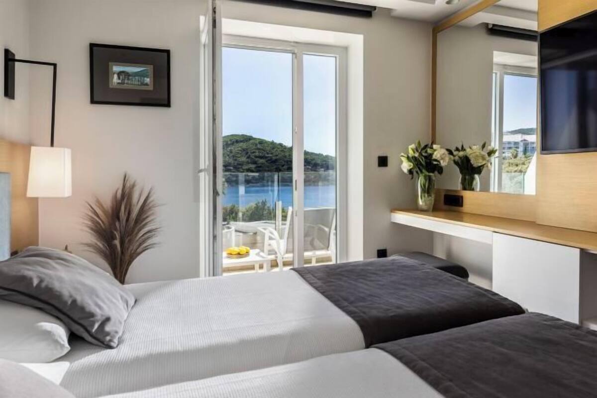 Un dormitorio con cama, mesita de noche, espejo y vista a un balcón con una silla y una mesa.