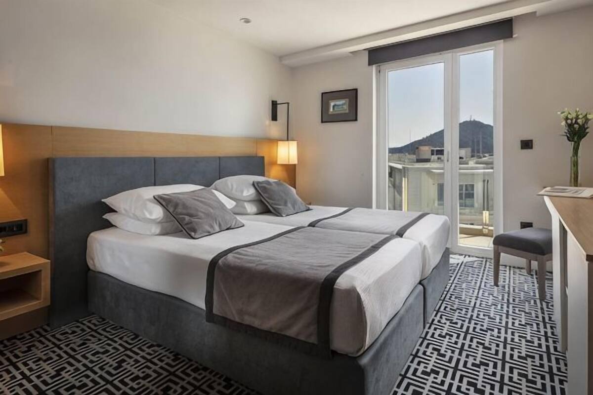 Habitación de hotel con cama doble, mesitas de noche, un escritorio y vista a un edificio a través de un ventanal grande.