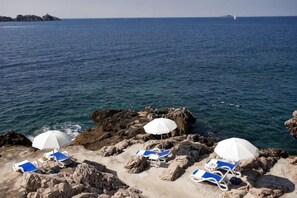 A coastal scene with rocky terrain, clear blue water, and lounge chairs with umbrellas. - Royal Neptun Hotel (Dubrovnik)