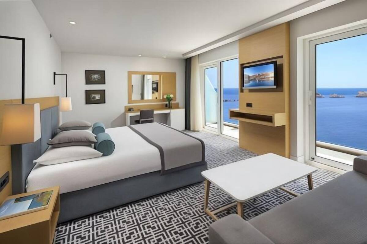 Habitación de hotel moderna con cama, escritorio, televisor y grandes ventanales con vista al mar.