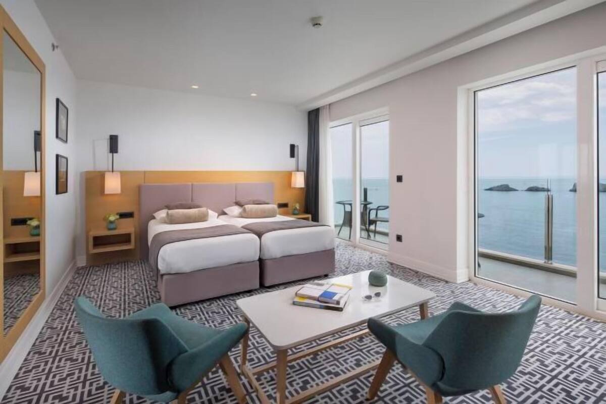 Una habitación de hotel moderna con una cama grande, una mesita pequeña, dos sillas y un balcón con vistas al mar.