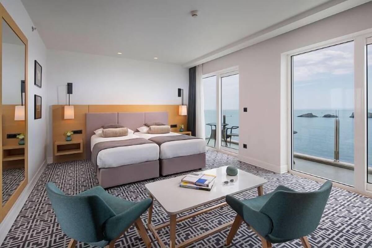 Habitación de hotel moderna con una cama grande, dos mesitas de noche, una pequeña mesa y vista al mar.