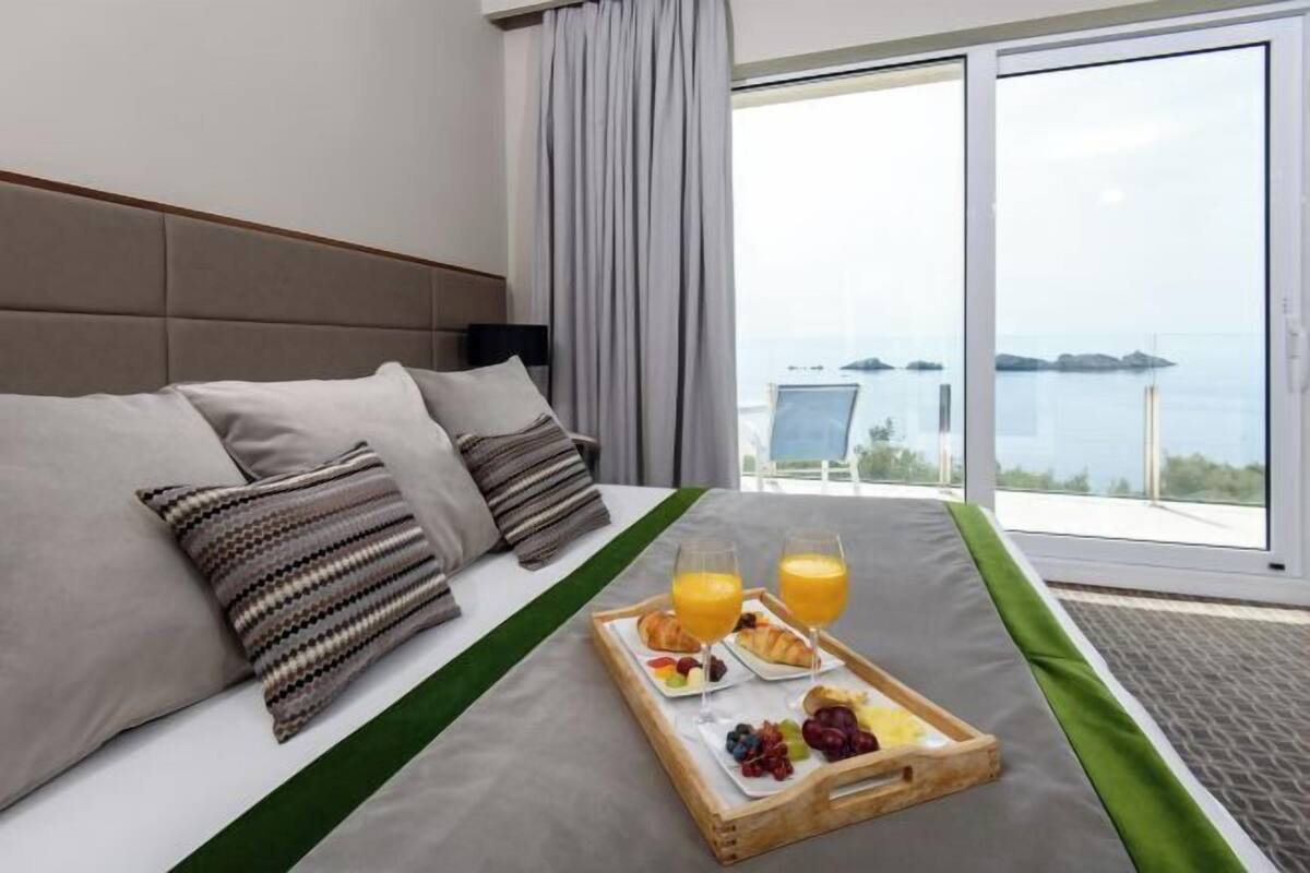Habitación de hotel con cama, bandeja de desayuno con cruasanes, fruta y jugo de naranja, y vista al mar.