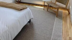 Une chambre d’hôtel avec un lit, deux fenêtres, une chaise, une petite table et un miroir.