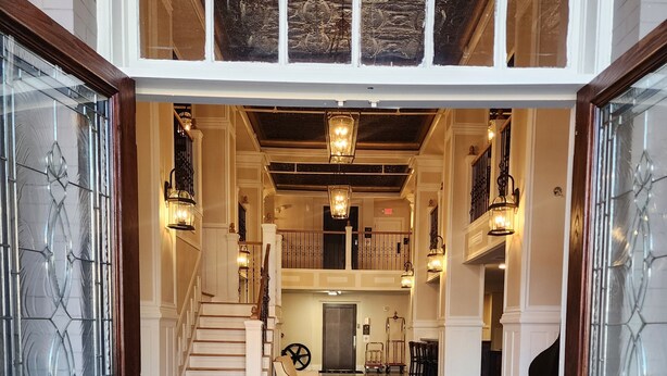 A grand entrance with a black doormat reading "HG" leading to a spacious foyer with a staircase, chandeliers, and a piano.