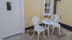 Een klein terras met twee witte metalen stoelen en een tafel, naast een geel gebouw met een witte deur en een raam.