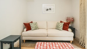 A living room with a beige sofa, patterned cushions, a small side table, a red patterned rug, a lamp, and a framed picture on the wall. - Cedar (Newbury)