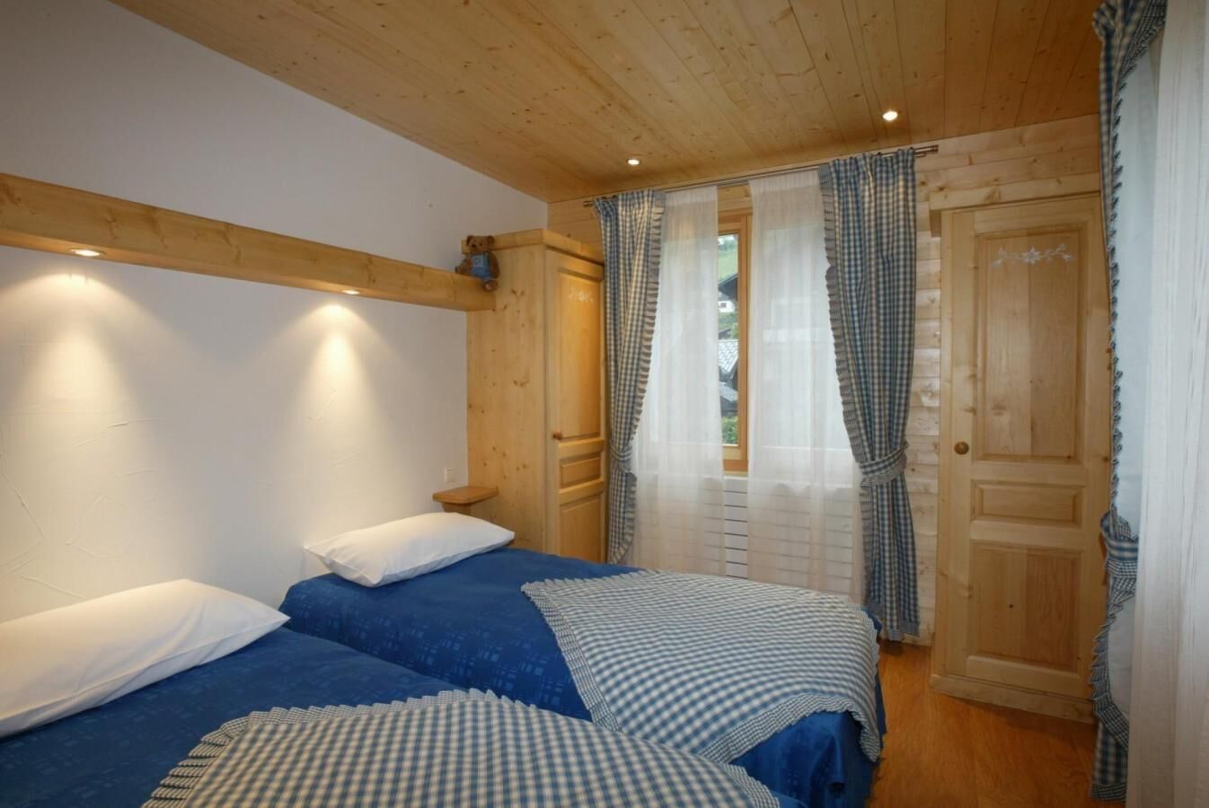 A bedroom with two beds, wooden ceiling beams, a wooden door, and a window with curtains.