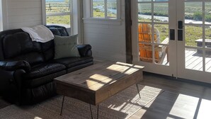 Amplia sala de estar con un sofá de cuero negro, una mesa de centro de madera y un tapete estampado. Grandes ventanales ofrecen vistas al exterior.