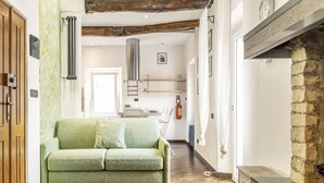 Una sala de estar con un sofá verde, vigas de madera, una chimenea de piedra y una cocina al fondo.