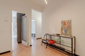 Un couloir avec une porte donnant sur une chambre, une console en verre surmontée d’une œuvre d’art colorée et une chaise.