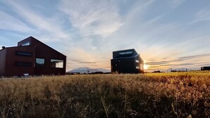 Un edificio moderno con techo plano y grandes ventanales, ubicado en un campo cubierto de hierba con montañas al fondo.