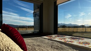 A bedroom with a large window, a bed with a patterned bedspread, and a view of a field and mountains. - Cabañas Julián Elias Patagónia (Puerto Natales)