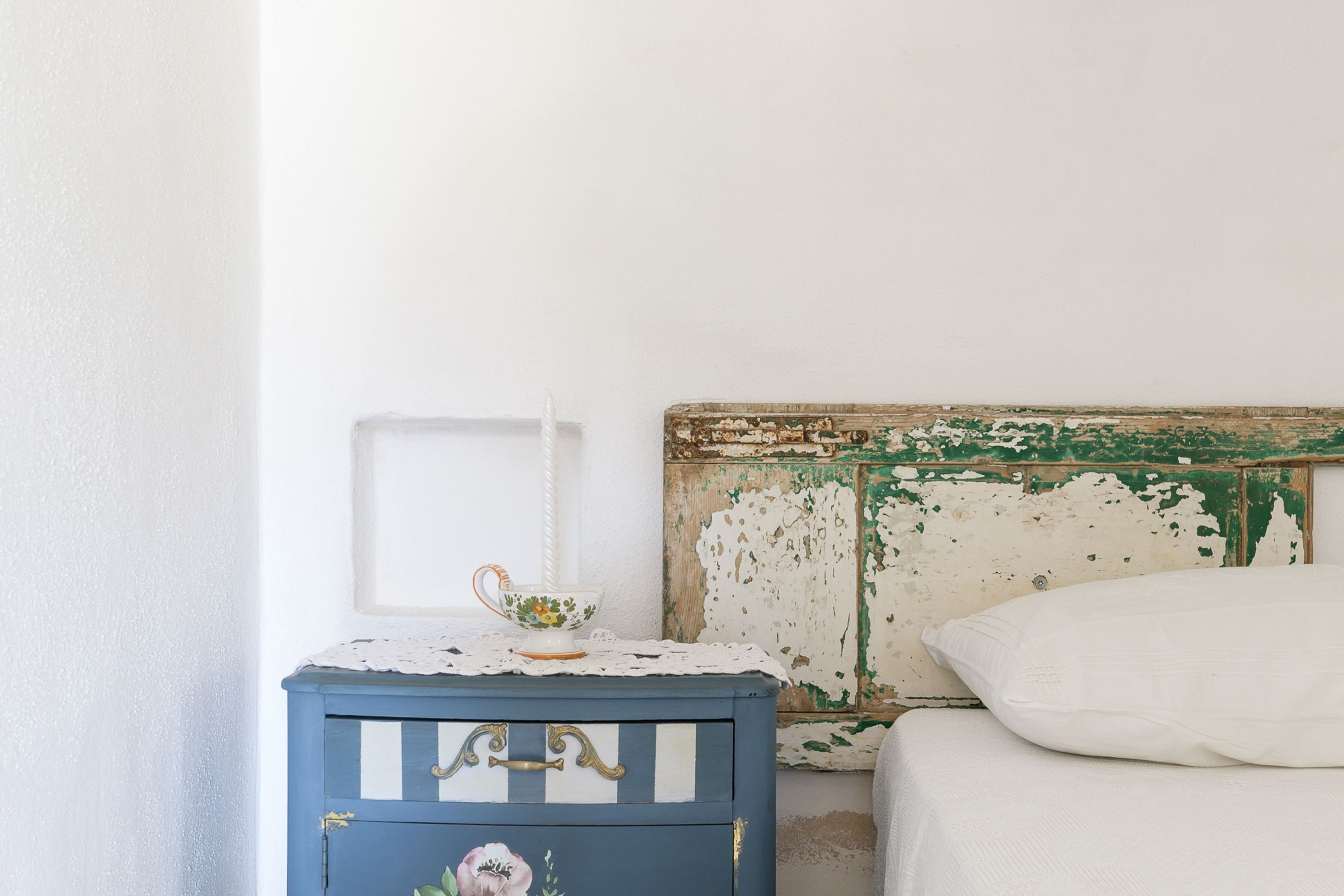 Une chambre à coucher avec un lit, une table de chevet ornée d’un objet décoratif et un mur dont la peinture s’écaille.