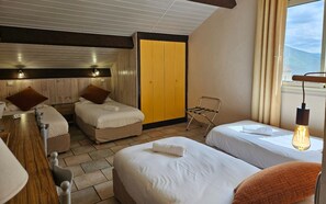 A hotel room with two beds, a wooden headboard, a yellow wardrobe, a window with a view, and a hanging light bulb. - Logis Hotel Le Mont Barral (Châtillon-en-Diois)