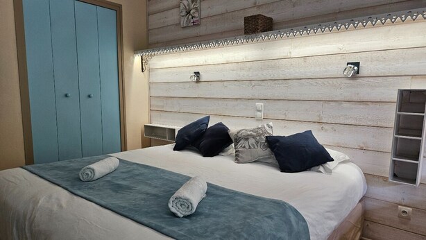 A bedroom with a wooden headboard, a bed with a white comforter, two rolled towels, and several pillows. - Logis Hotel Le Mont Barral (Châtillon-en-Diois)