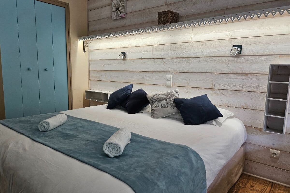 A bedroom with a wooden headboard, a bed with a white comforter, two rolled towels, and several pillows.