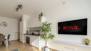Moderni olohuone, jossa on seinälle asennettu taulutelevisio, ruokailutila pöydän ja tuolien kera, valkoisilla kaapistoilla varustettu keittiö sekä ruukkukasvi.