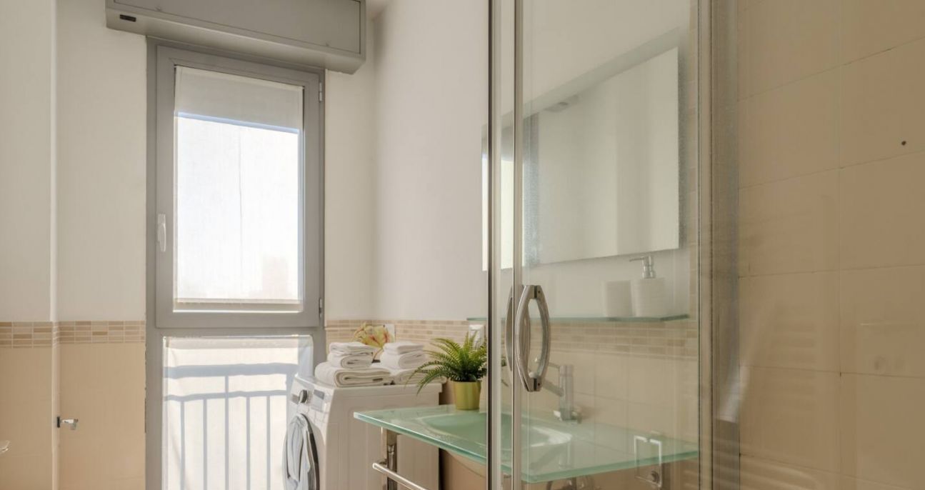 Un baño con cabina de ducha de vidrio, lavamanos de vidrio, toallero y ventana con persianas.